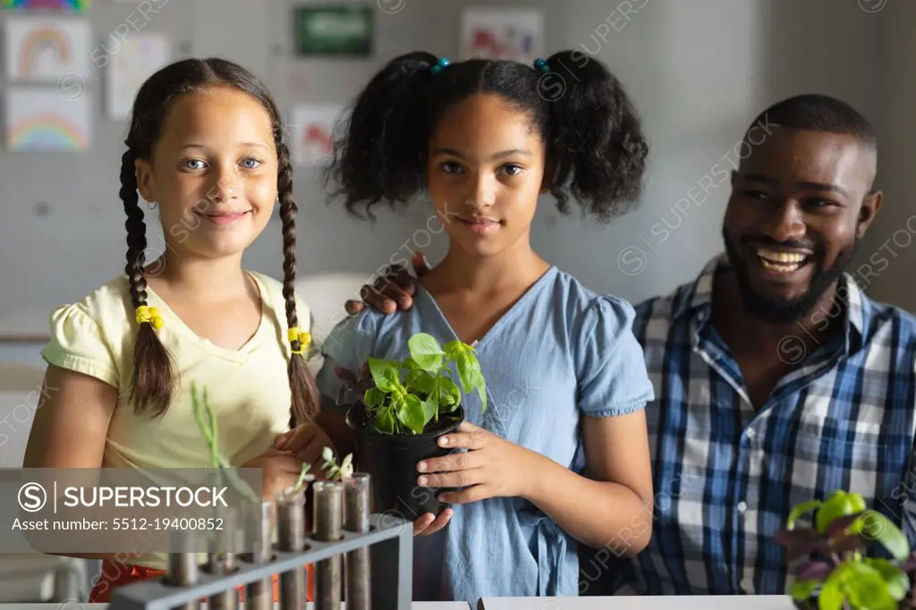 Portrait of biracial elementary schoolgirls with plant standing by african american young teacher. unaltered, education, childhood, teaching, science, stem and school concept.