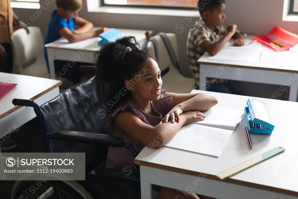 Smiling biracial elementary schoolgirl sitting on wheelchair at desk in classroom. unaltered, education, childhood, learning, disability, physical disability and school concept.