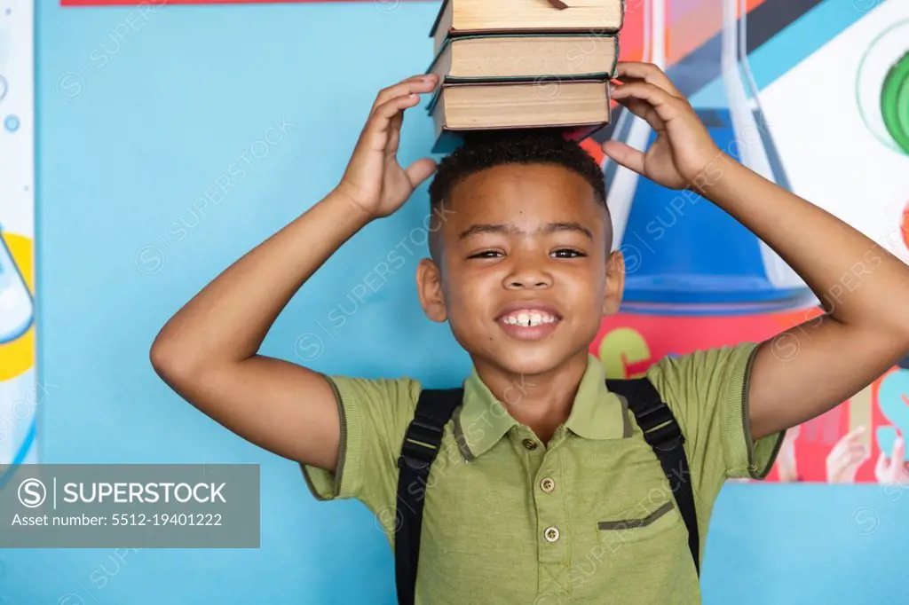Portrait of smiling african american elementary schoolboy stacking books on head against wall. unaltered, education, childhood and school concept.