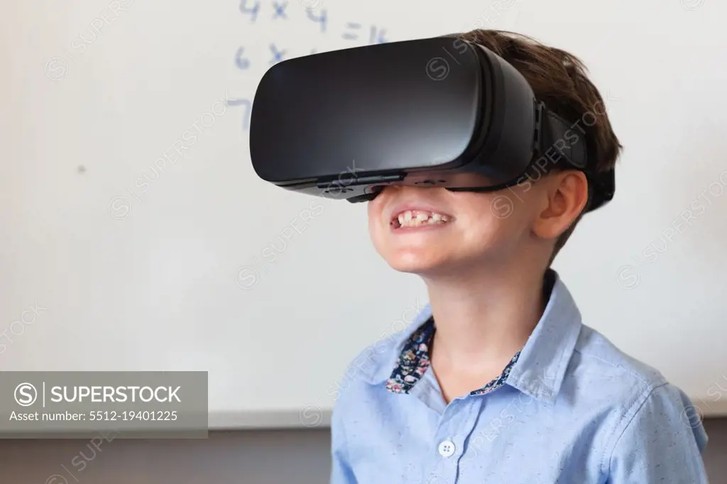 Smiling caucasian elementary schoolboy wearing vr glasses against whiteboard in classroom. unaltered, education, virtual reality simulator, technology and school concept.