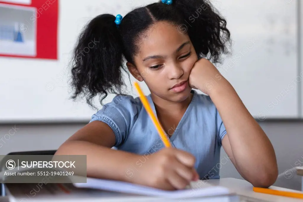 Biracial elementary schoolgirl writing on book at desk in classroom. unaltered, education, learning, studying, concentration and school concept.