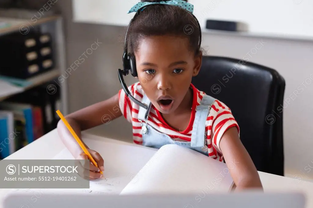 African american elementary schoolgirl with mouth open looking at laptop while studying at desk. unaltered, education, listening, studying, concentration, wireless technology and school concept.