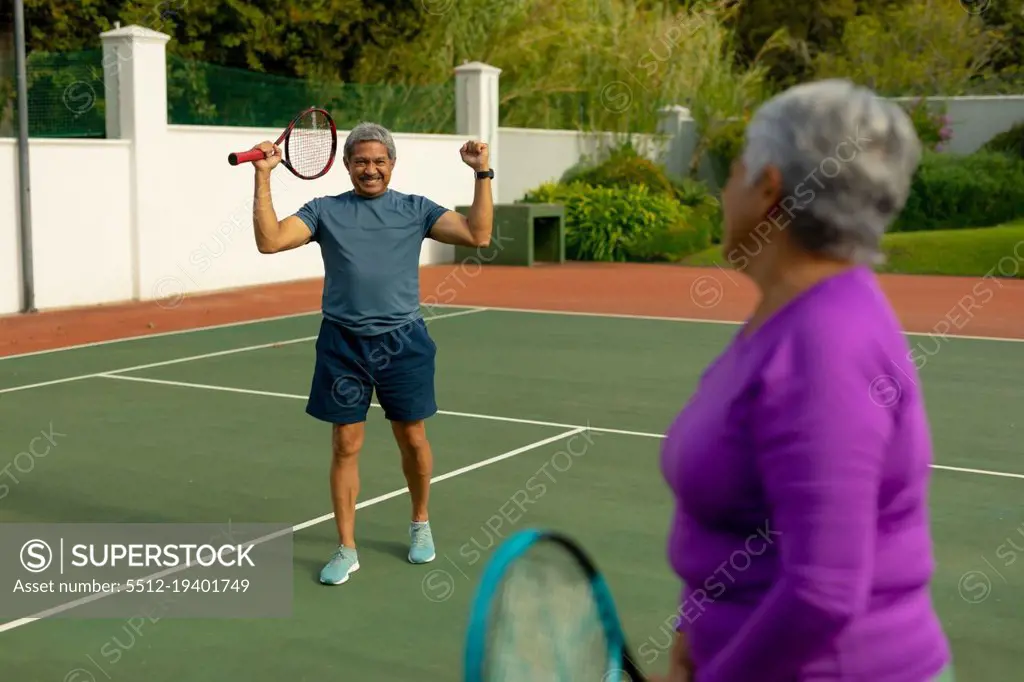 Biracial senior woman looking at cheerful senior man holding racket and gesturing in tennis court. unaltered, sport, competition, togetherness, achievement, retirement, healthy lifestyle concept.