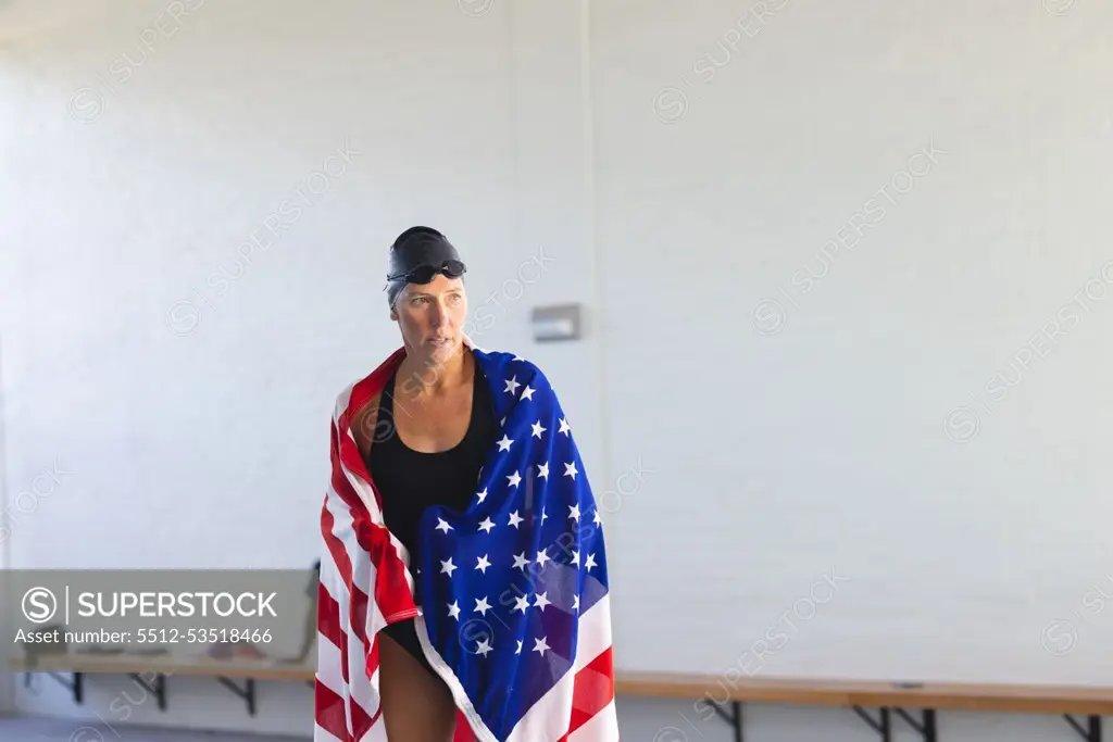 Caucasian female athlete swimmer wrapped in an American flag, with copy space. She stands confidently in a gym setting, suggesting athletic pride or patriotism.