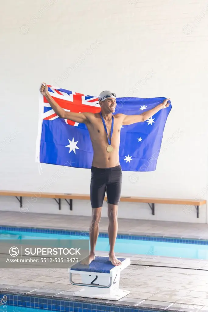 Young biracial male swimmer celebrates victory at a swimming pool with the Australian flag. Holding the Australian flag, the athlete stands proud with a medal, symbolizing triumph.