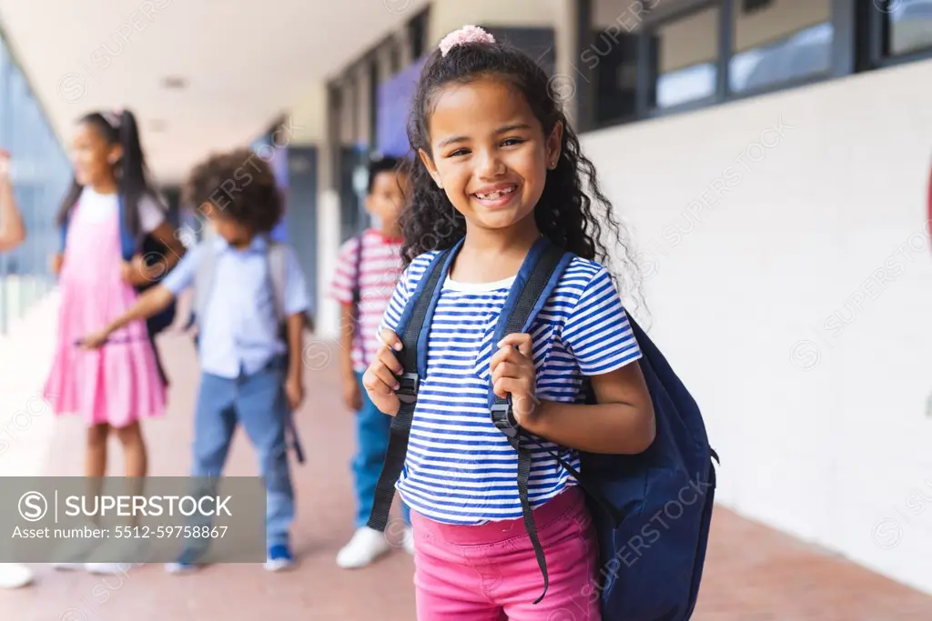 Students, including biracial girl and African American boy, smiling with backpacks. Standing outside school building, ready for a day of learning and fun, unaltered