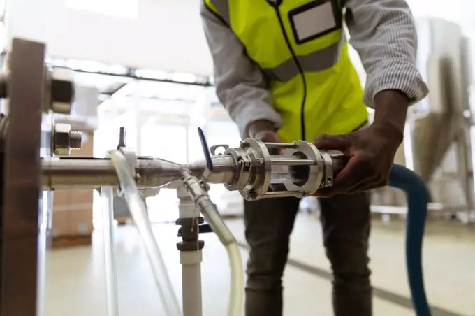 Mid section view of an African American man working at a microbrewery, wearing a high visibility vest, inspecting a tank pipe.