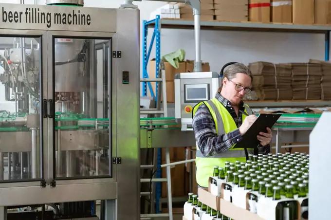 Caucasian man wearing high visibility vest, working in a microbrewery, holding a file and writing data while checking bottles of beer prepared for delivery..