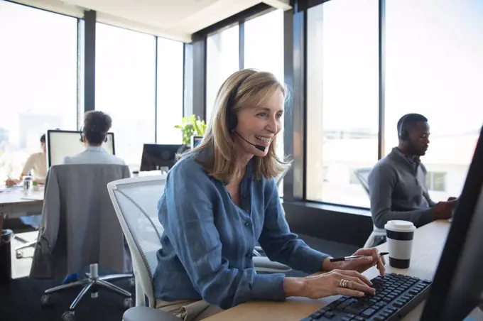 A Caucasian businesswoman working in a modern office, sitting at a desk, using a laptop computer, wearing headset and talking, with her colleagues working in the background