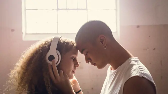 Side view of a young Hispanic-American and a young Caucasian woman wearing headphones with eyes closed and foreheads meeting inside an empty warehouse