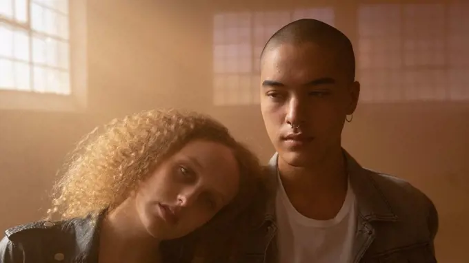 Front view of a young Caucasian female with curly hair looking intently at the camera while resting head on the shoulder of a young Hispanic-American man inside an empty warehouse