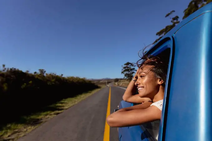 Side view of a young mixed race woman leaning out of the front passenger side window of a pick-up truck smiling, as it drives down the highway on a road trip 