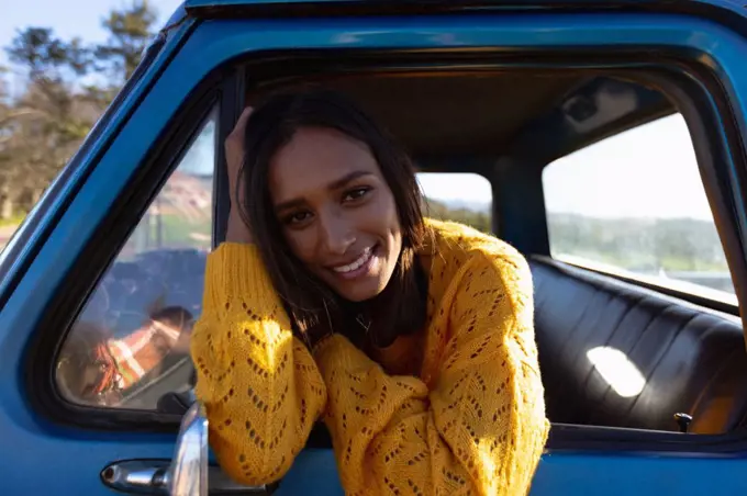 Portrait close up of a young mixed race woman sitting in the front passenger seat of a pick-up truck, leaning out of the side window smiling to camera during a road trip