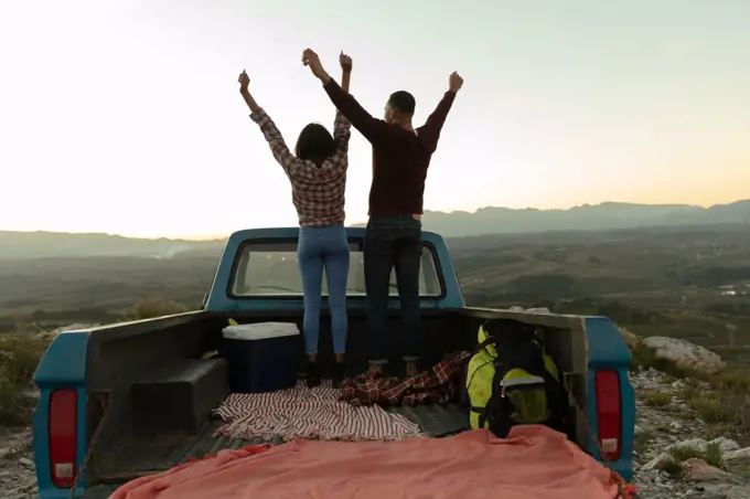 Back view of a young mixed race couple standing on the back of their pick-up truck with their arms raised enjoying the view at sundown during a stop off on a road trip.