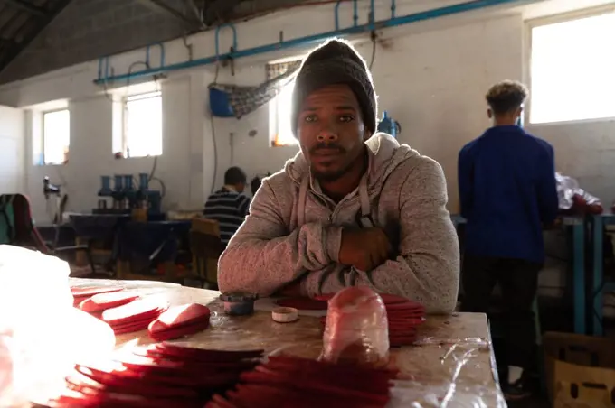 Portrait close up of a young African American man sitting at a workbench in a factory makimg cricket balls, looking to camera and smiling. In the background colleagues are working on other parts of the production line. They are working in a clothing factory.