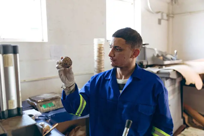 Front view close up of a young mixed race man wearing gloves and overalls holding the core of a ball and checking it at a factory making cricket balls, with equipment and materials visible in the background. They are working in a clothing factory.