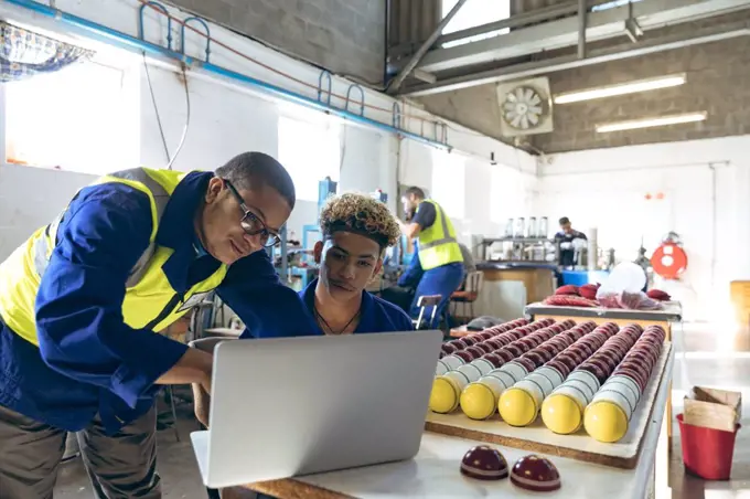 Front view close up of a young mixed race male manager standing and looking at a laptop with a young mixed race male worker sitting at a workbench with finished cricket balls at a cricket ball factory. They are working in a clothing factory.