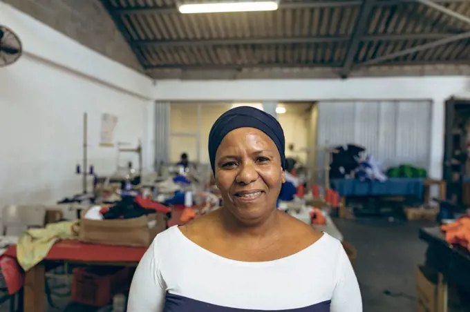 Portrait close up of a middle aged mixed race woman in a brightly lit sports clothing factory, looking to camera and smiling. They are working in a clothing factory.