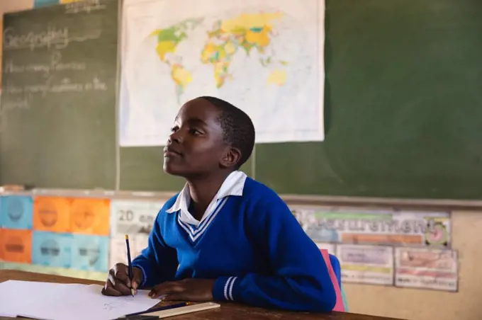 Side view close up of a young African schoolboy sitting at a desk looking up while writing in his note book and listening attentively during a lesson in a township elementary school classroom