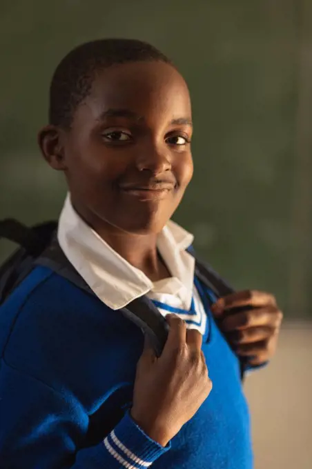 Portrait close up of a young African schoolboy wearing his school uniform and schoolbag, looking straight to camera smiling, at a township elementary school