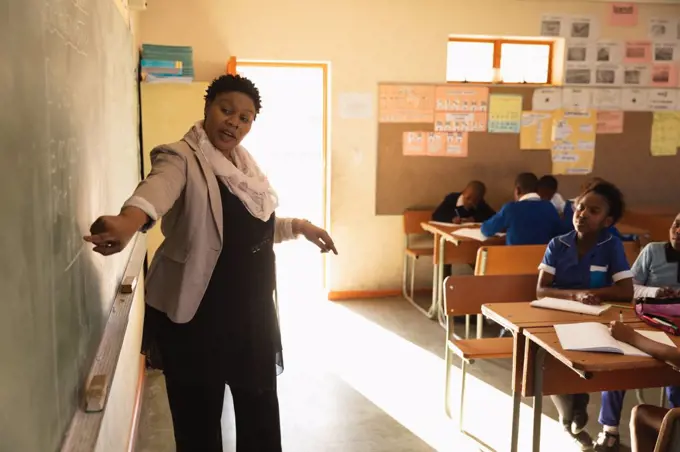 Front view close up of a middle aged African female school teacher standing at the front of the class pointing at the blackboard with pupils watching from their desks during a lesson in a township elementary school classroom