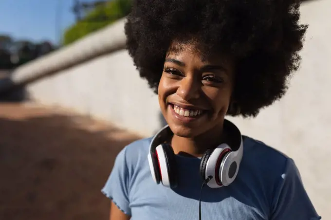 Portrait close up of a smiling young mixed race woman wearing headphones around her neck in a sunny urban park