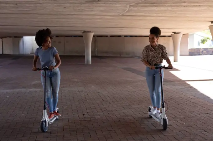 Front view of two young adult mixed race sisters riding on electric scooters in an urban park, looking at each other smiling