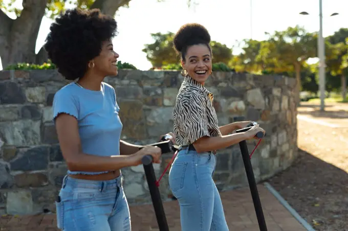 Side view close up of two young adult mixed race sisters riding on electric scooters in an urban park, looking at each other smiling