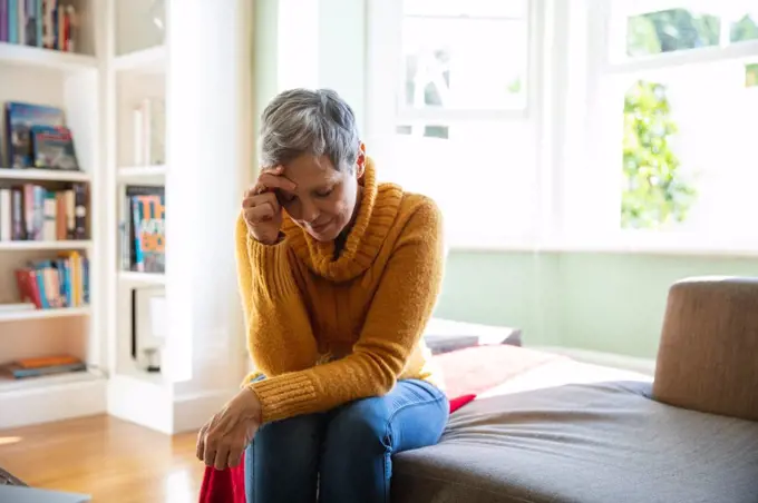 Front view close up of a mature Caucasian woman with short grey hair sitting at home in her living room looking down with her head leaning on her hand, a sunlit window in the background