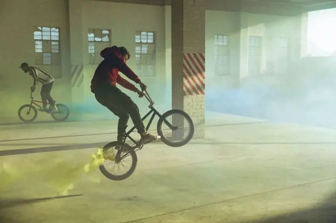 Side view of two young Caucasian men riding and doing tricks on BMX bikes with yellow and blue smoke grenades attached to them, in an abandoned warehouse