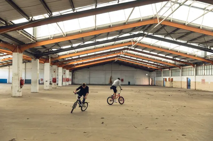 Front view of two young Caucasian men facing opposite directions riding BMX bikes while practicing tricks in an abandoned warehouse