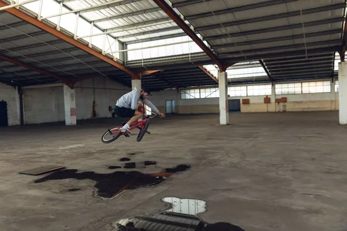 Side view of a young Caucasian man riding a BMX bike, jumping off the ground and turning the handlebars, while practicing tricks in an abandoned warehouse