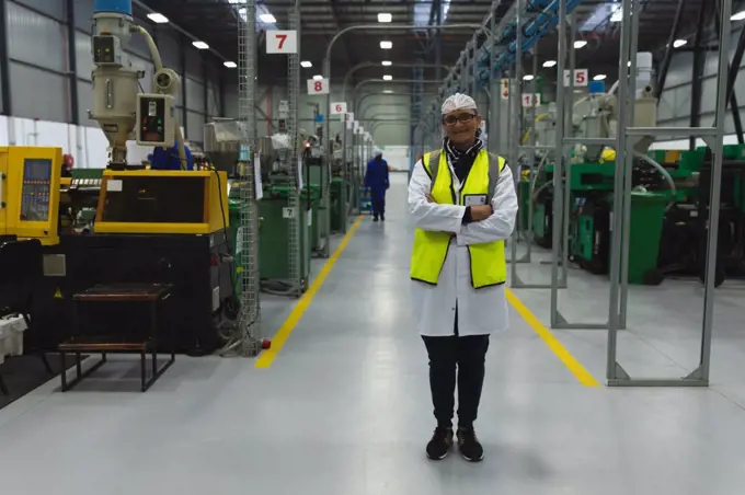 Portrait of a middle aged Caucasian woman wearing glasses and workwear standing between rows of equipment smiling in a warehouse at a processing plant, another worker visible in the background 