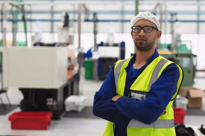 Portrait close up of a middle aged mixed race male factory worker wearing glasses and workwear looking to camera with arms crossed in a warehouse at a processing plant