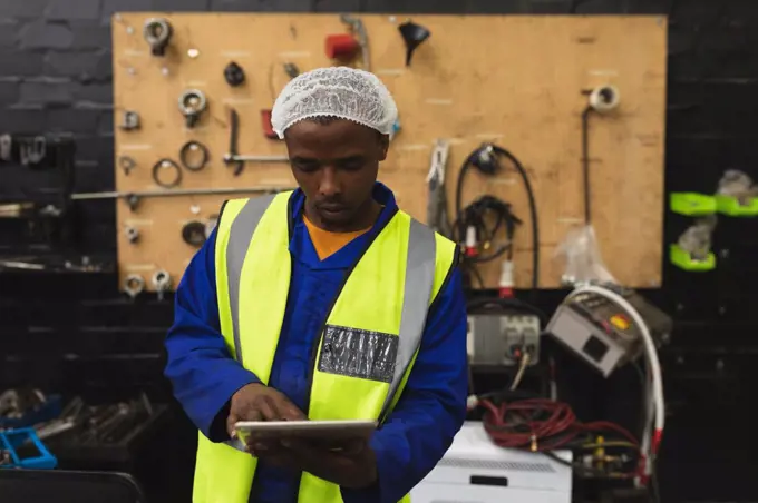 Front view close up of a young African American male factory worker using a tablet computer in the machine shop at a processing plant, with equipment and tools in the background