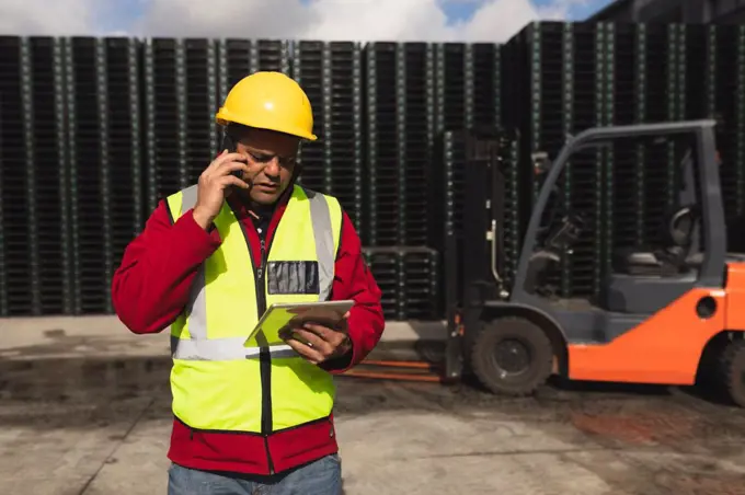 Front view close up of a middle aged Caucasian male factory worker on the phone using a tablet computer beside a forklift truck and stacks of pallets outside a warehouse at a factory 