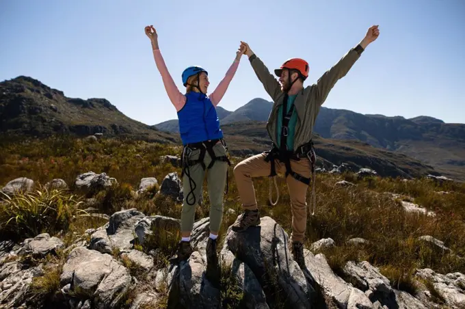 Front view of Caucasian couple enjoying time in nature, wearing zip lining equipment, holding hands with arms in the air, smiling at each other on a sunny day in mountains