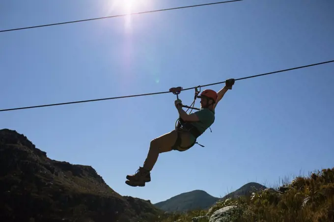 Side view of Caucasian man enjoying time in nature, zip lining on a sunny day in mountains. Fun adventure vactation weekend.