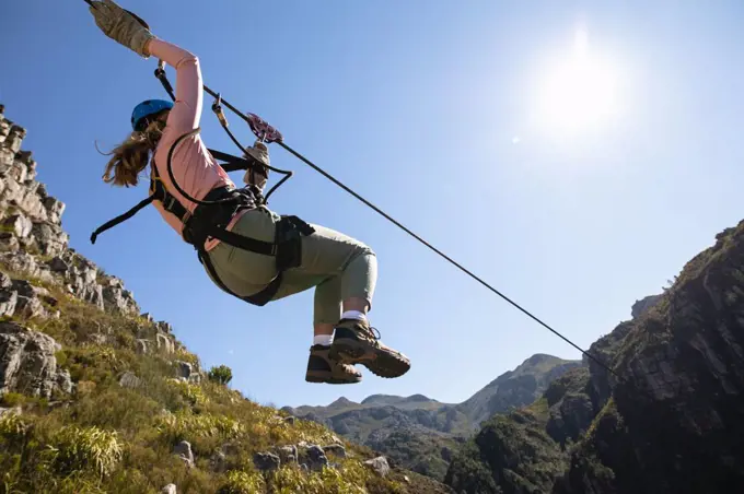 Low angle rear view of Caucasian woman enjoying time in nature, zip lining  on a sunny day in mountains. Fun adventure vactation weekend.