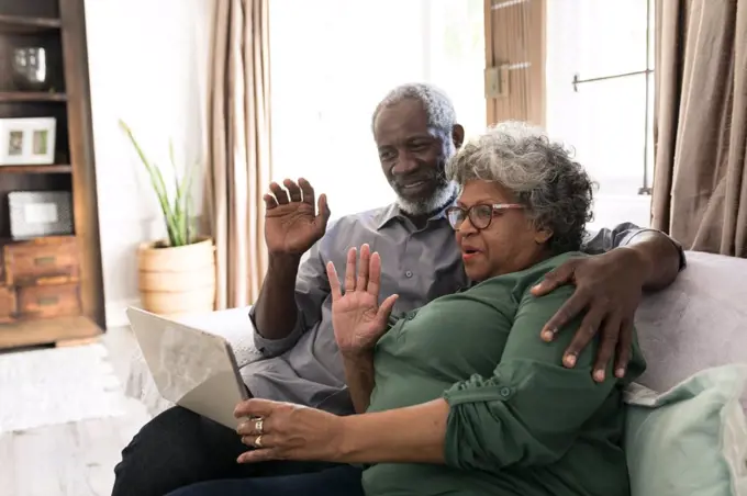 A senior African American couple spending time at home together, social distancing and self isolation in quarantine lockdown during coronavirus covid 19 epidemic, using a tablet, making a video call to friends or relatives, waving