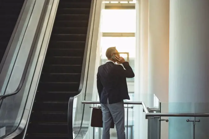 Rear view of businessman with briefcase talking on the phone near the escalator in office