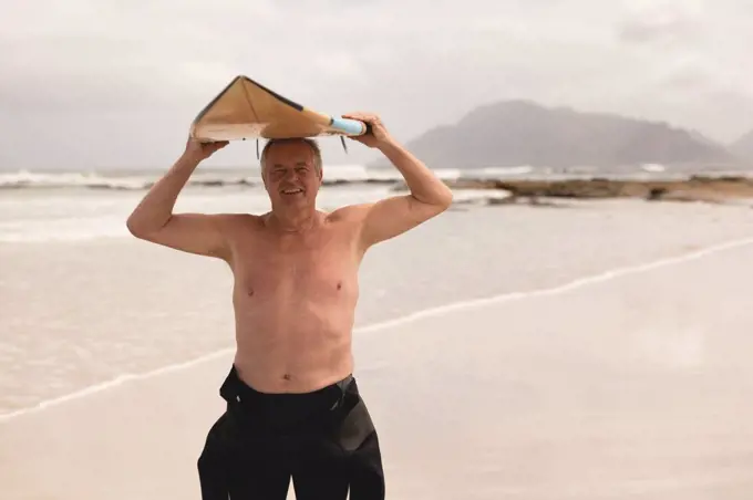 Front view of active senior man carrying surfboard on his head at beach against mountains background