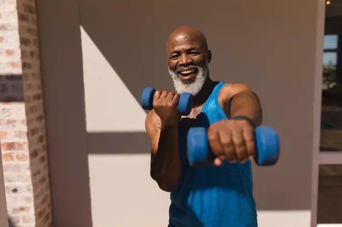 Front view of active senior African American man training arms with dumbbells in the backyard of home