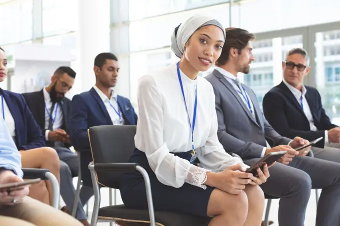 Front view of mixed race businesswoman with digital tablet looking at camera in seminar. businessmen interacting behinf her