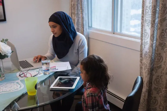 Side view of mixed race mother wearing hijab using laptop while daughter looking at digital tablet at home. They are sitting around a table in living room