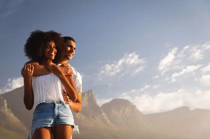 Low angle view of African-american couple standing together in romantic mood near sea side. They are smiling and looking away in the same direction with mountains on background