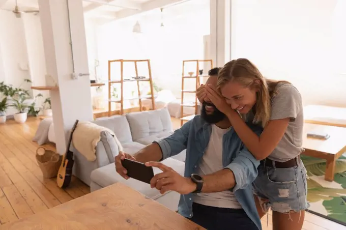 Front view of african-american Man taking selfie while caucasian woman closing his eyes at home