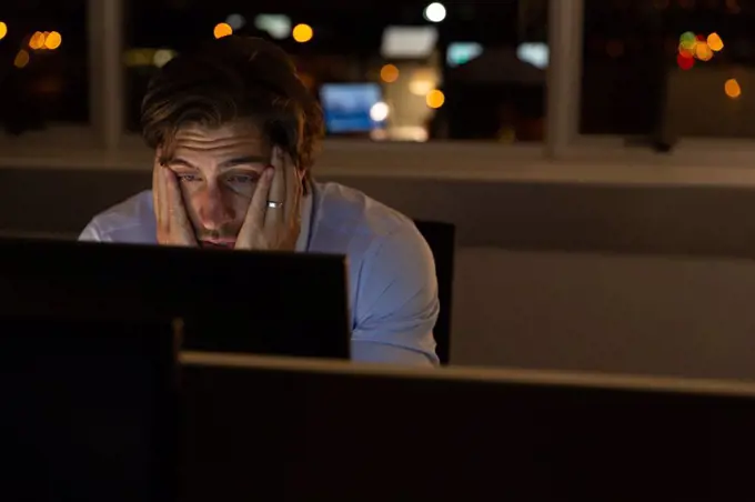 Front view of tired young Caucasian male executive with hands on face working at desk in a modern office. He is working late