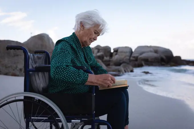 Side view of a disabled active Caucasian senior woman reading a book on a wheelchair next to the water on the beach