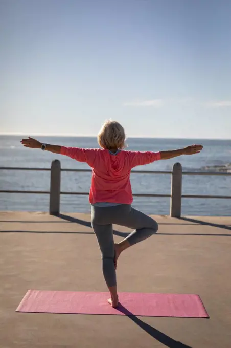 Rear view of an active senior woman performing yoga on a fitness map on a promenade in front of the sea